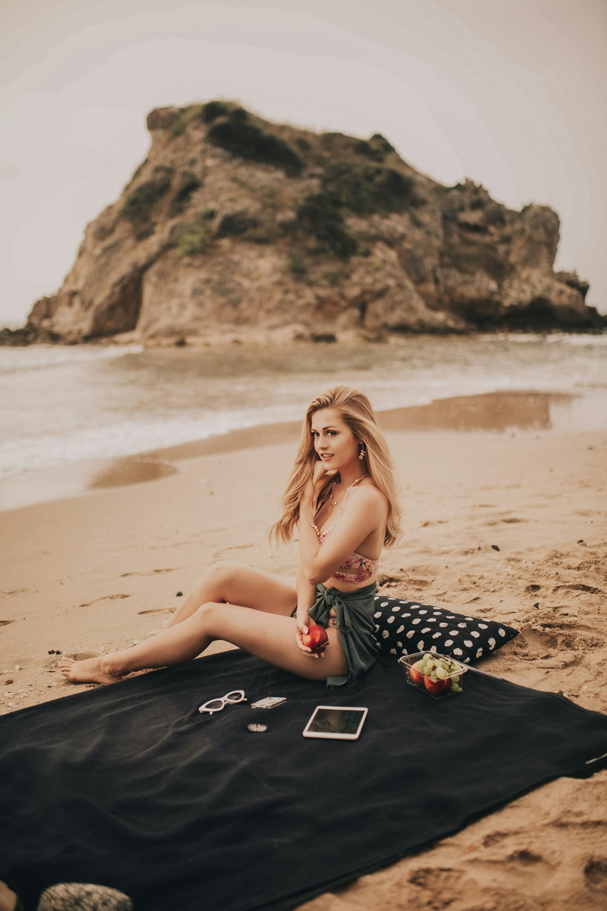 Девушка сидит на черном покрывале на пляже.
